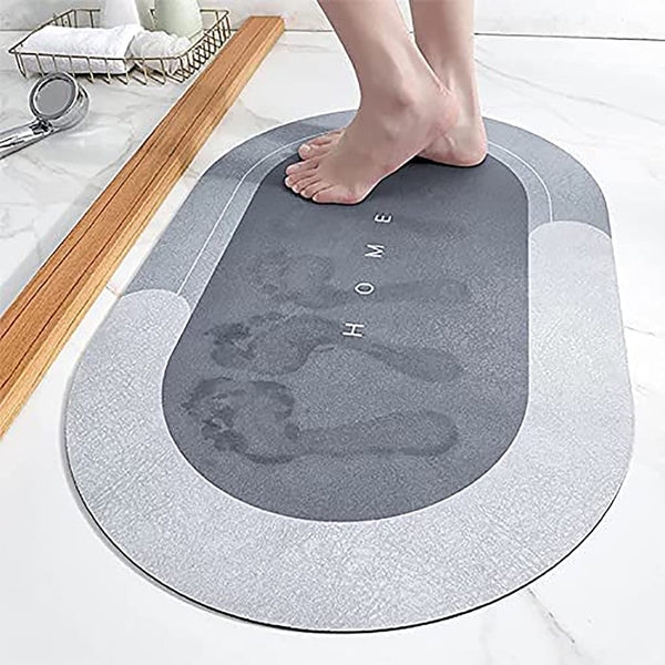 A gray oval-shaped bath mat with 'HOME' text on it, placed on a tiled floor with a person's foot for scale.